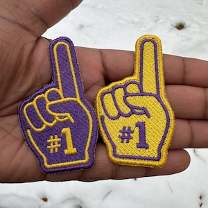 May include: Two embroidered foam finger patches in purple and yellow, each with a gold outline and the number "#1". The patches are in the shape of a hand with the index finger pointing up.