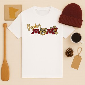 May include: White t-shirt with a "Gopher Mom" graphic in gold, maroon, and tan. The design features a cartoon gopher mascot. A maroon knit beanie, a coffee cup, and a pinecone are also visible.
