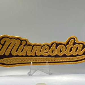 May include: An embroidered patch featuring the word Minnesota in a cursive script font. The lettering is rendered in a bright yellow thread with a dark brown background and a thick yellow border, creating a bold and distinct visual contrast.