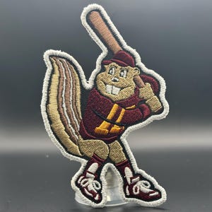 May include: Embroidered patch featuring a cartoon squirrel in a baseball uniform, holding a bat. The squirrel is wearing a maroon jersey with a gold "A" and a matching cap. The patch has a white border and is on a clear plastic stand.