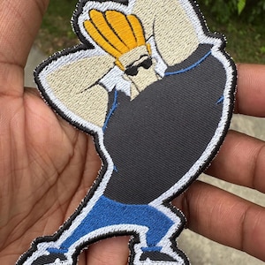 May include: Embroidered patch of a cartoon character with yellow hair, sunglasses, and a black shirt. The patch has a white border and blue trousers. The character is in a dynamic pose, with arms raised.