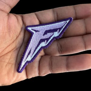 May include: Embroidered patch in a stylized lightning bolt shape. The patch features a purple design with white and light purple accents, outlined with a purple border. The patch is held in a hand.