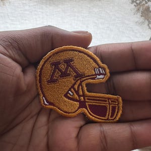 Può includere: Toppa ricamata di un casco da football americano in oro e bordeaux. Il casco presenta la lettera "M" in bordeaux sul lato. La toppa ha un bordo bianco.