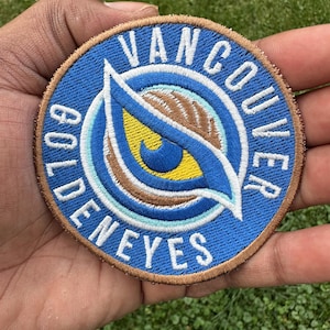 Puede incluir: Un parche bordado circular con fondo azul y el texto VANCOUVER GOLDENEYES en blanco. El centro muestra un gráfico de ojo estilizado con un iris amarillo, una pupila azul y un elemento circular texturizado de color marrón detrás.