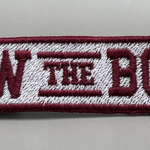 May include: Embroidered patch with the words "ROW THE BOAT" in maroon thread on a white background. The patch has a maroon border and is rectangular in shape. The text is in a bold, sans-serif font.