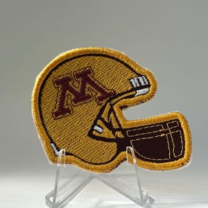 May include: An embroidered patch shaped like a football helmet in gold, maroon, and white. The helmet features a maroon letter M on the side and a white face mask. The patch has a thick gold border and is displayed on a clear plastic stand.