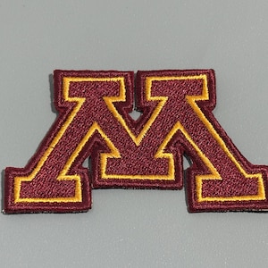 May include: Embroidered patch featuring a maroon "M" with gold trim. The patch has a stitched border and is likely designed for application on clothing or accessories. The design is a bold, collegiate-style letter.