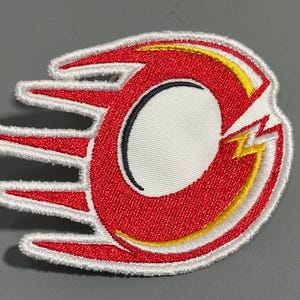 May include: Embroidered patch featuring a stylized red and white design with yellow accents. The patch has a circular shape with a lightning bolt and a white border. The design is on a white background.