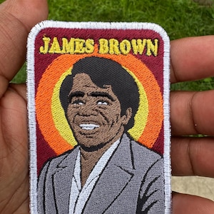 May include: Embroidered patch featuring a portrait of James Brown. The patch has a rectangular shape with a white border. The image of James Brown is set against a backdrop of concentric orange, yellow, and red circles, with the text "JAMES BROWN" at the top.