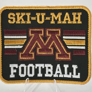May include: A rectangular embroidered patch with a black background and a gold border. The text "SKI-U-MAH" is stitched in gold at the top, and "FOOTBALL" is in white at the bottom. A large maroon "M" logo sits in the center with horizontal stripes.