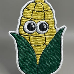 Op de afbeelding: Geborduurde patch met een cartoon maïskolf met grote ogen. De maïs is geel, met groene bladeren en een witte rand. Een speels ontwerp van een veel voorkomende groente, voor kleding of accessoires.