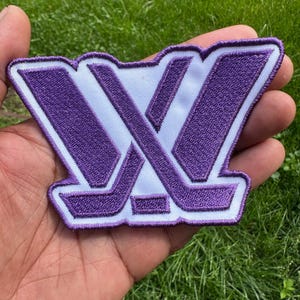 May include: A purple and white embroidered patch featuring a stylized letter W design. The emblem is constructed with thick purple thread forming the letter shape, outlined and filled with a white fabric base, creating a bold, high-contrast graphic.