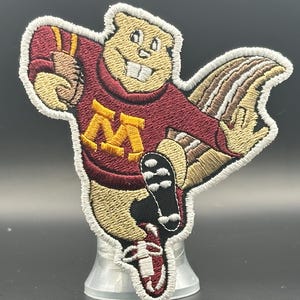 May include: Embroidered patch featuring a cartoon gopher in a maroon jersey with a gold "M" logo. The gopher is in a running pose, with a detailed tail and a white outline. The patch is on a clear plastic stand.