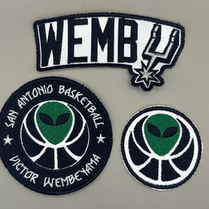 Könnte beinhalten: Drei gestickte Aufnäher mit Basketball-Motiv. Ein Aufnäher trägt die Aufschrift "WEMB" mit einem Blitzdesign. Zwei runde Aufnäher zeigen einen Alienkopf über einem Basketball mit dem Text "SAN ANTONIO BASKETBALL" und "VICTOR WEMBEYAMA".