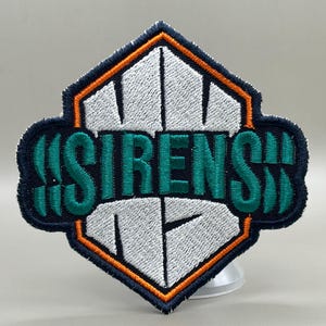 May include: Embroidered patch with the word "SIRENS" in teal, set against a white and navy blue background. The patch has an orange border and a diamond-shaped design. The patch is likely for clothing or accessories.