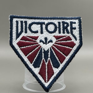 May include: Embroidered patch featuring the word "VICTOIRE" in white, navy blue, and burgundy. The patch has a white border and a decorative design. The patch is shield-shaped.