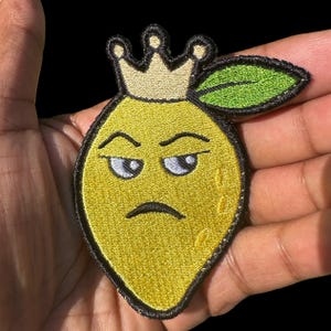 Puede incluir: Parche bordado con un limón amarillo coronado, con una hoja verde y una expresión de mal humor. El parche tiene un borde negro, adecuado para ropa o accesorios.