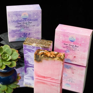 May include: Two bars of handcrafted soap, one lavender and one rose, with their packaging boxes. The lavender soap bar is purple and the rose soap bar is pink. Both bars have a layer of dried flowers and herbs on top. The boxes are decorated with butterflies and stars.