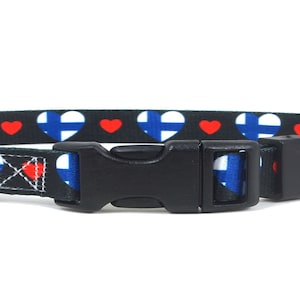 May include: A black dog collar with a repeating pattern of red hearts and blue and white Finnish flags. The collar has a black plastic buckle and a silver metal D-ring.
