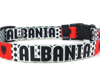May include: A black and white dog collar with the word "ALBANIA" printed on it. The collar has a red and black Albanian flag on each side.