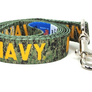 May include: A green and yellow camouflage dog leash with the word "NAVY" printed in yellow. The leash has a silver metal clip.