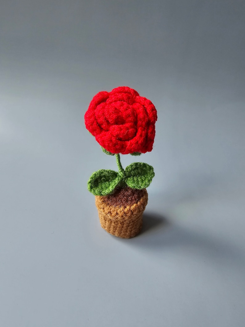 May include: A crocheted red rose in a brown pot with green leaves.
