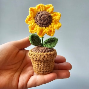May include: A crocheted sunflower potted plant with brown and yellow petals, green leaves, and a brown pot.