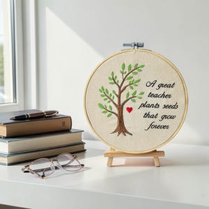 Peut inclure: Broderie d'arbre dans un cercle en bois avec le texte "A great teacher plants seeds that grow forever". L'œuvre présente un arbre brun avec des feuilles vertes et un cœur rouge, sur un petit chevalet en bois.