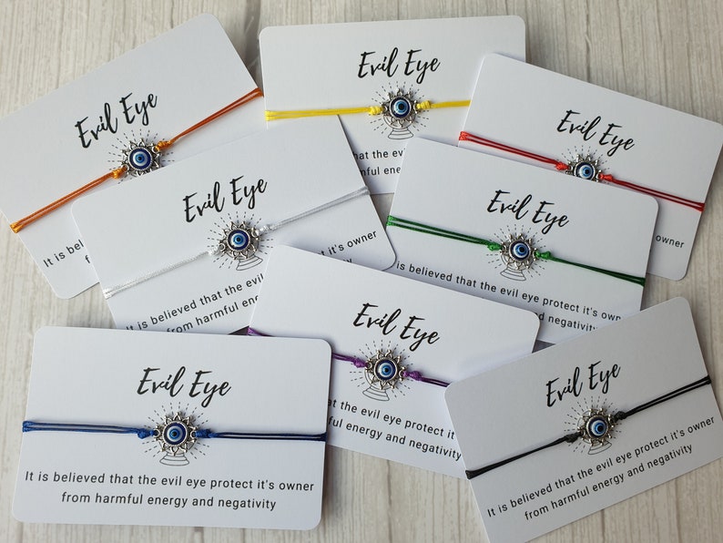 May include: Assortment of colorful string bracelets with silver evil eye charms. Each bracelet is accompanied by a card with the text "Evil Eye" and a description of the evil eye's protective properties.