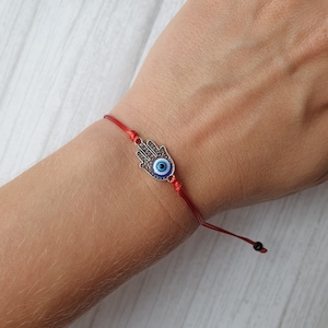 May include: Red string bracelet with a silver charm featuring a hamsa hand with a blue evil eye.