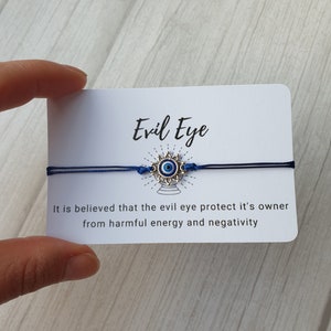 May include: A blue string bracelet with a silver charm featuring a blue evil eye. The card reads "Evil Eye" and "It is believed that the evil eye protect it's owner from harmful energy and negativity."