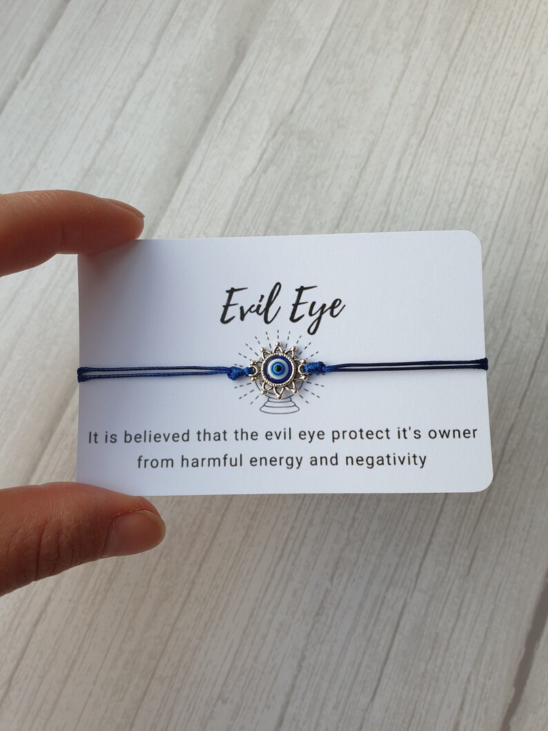 May include: A blue string bracelet with a silver charm featuring a blue evil eye. The card reads "Evil Eye" and "It is believed that the evil eye protect it's owner from harmful energy and negativity."