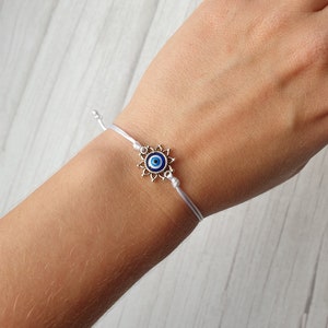 May include: White string bracelet with a silver charm featuring a blue evil eye design.