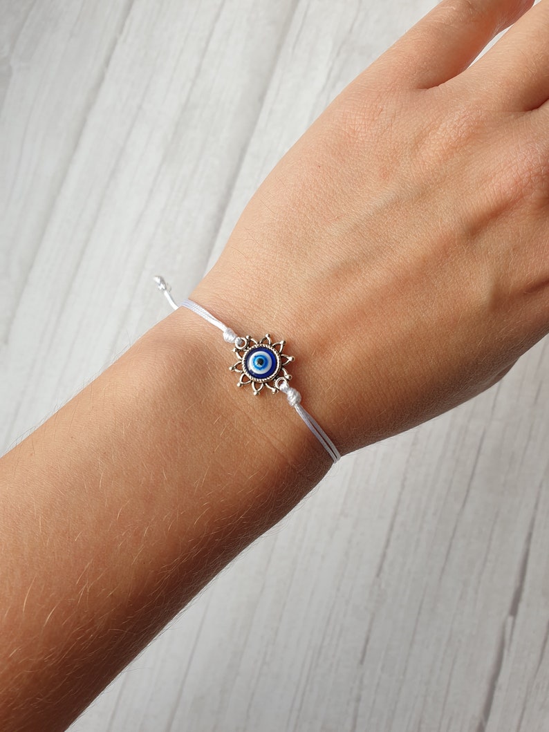 May include: White string bracelet with a silver charm featuring a blue evil eye design.