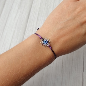 May include: A purple string bracelet with a silver charm featuring a blue evil eye.