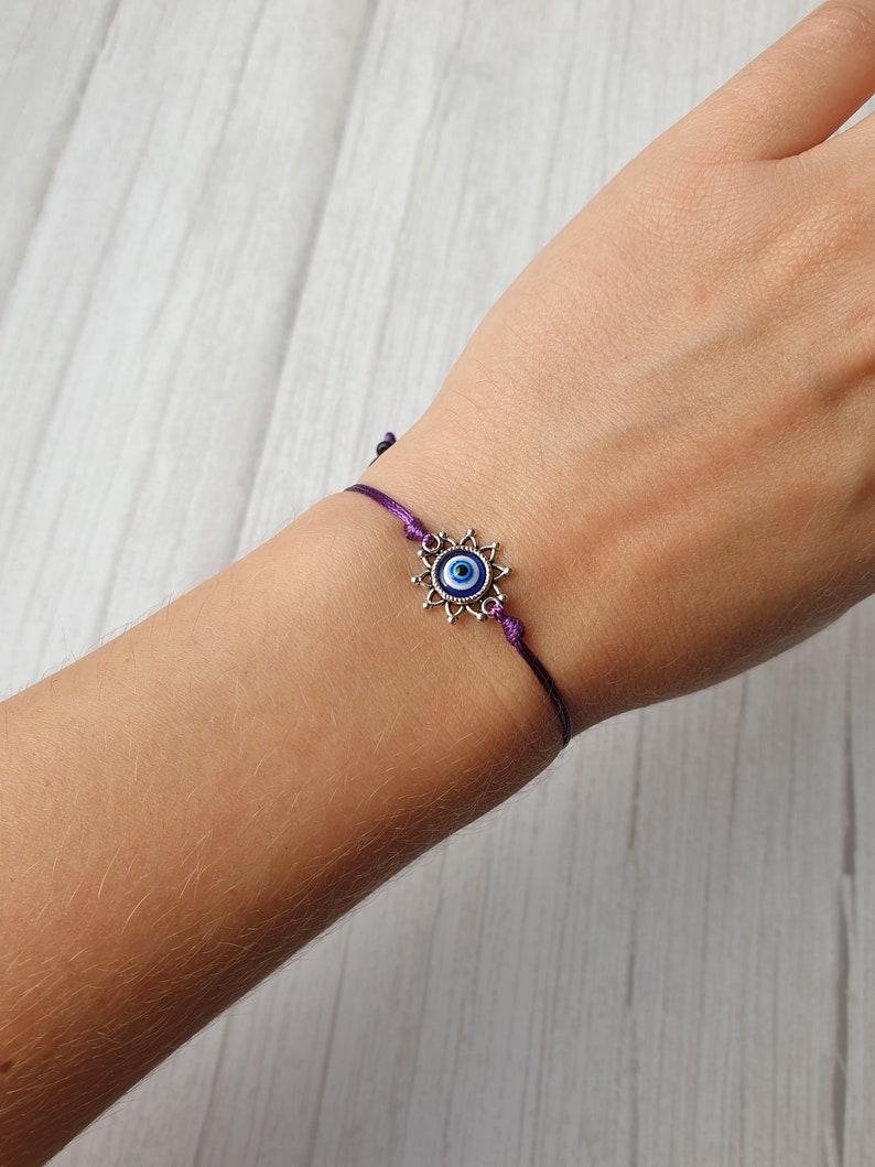 May include: A purple string bracelet with a silver charm featuring a blue evil eye.