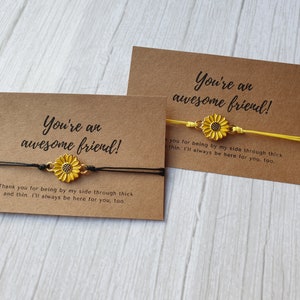 May include: Two brown cards with the text "You're an awesome friend!" and a sunflower charm bracelet. The bracelet on the left card has a black string and the bracelet on the right card has a yellow string.