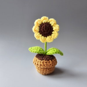 May include: A crocheted sunflower with a brown center and yellow petals, in a brown pot with green leaves.