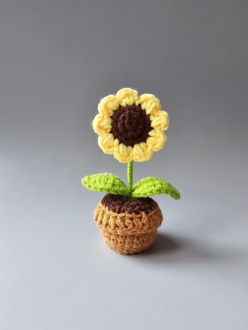 May include: A crocheted sunflower with a brown center and yellow petals, in a brown pot with green leaves.