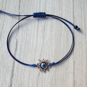 May include: A blue string bracelet with a silver charm featuring a blue evil eye. The charm has a starburst design around the eye.