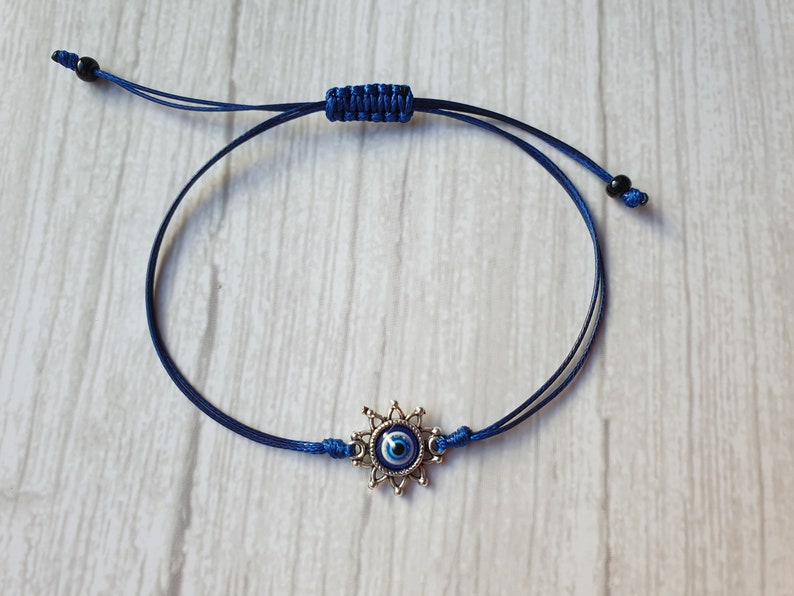 May include: A blue string bracelet with a silver charm featuring a blue evil eye. The charm has a starburst design around the eye.