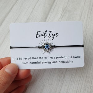 May include: A black string bracelet with a silver charm featuring a blue evil eye. The card reads "Evil Eye" and "It is believed that the evil eye protect it's owner from harmful energy and negativity."