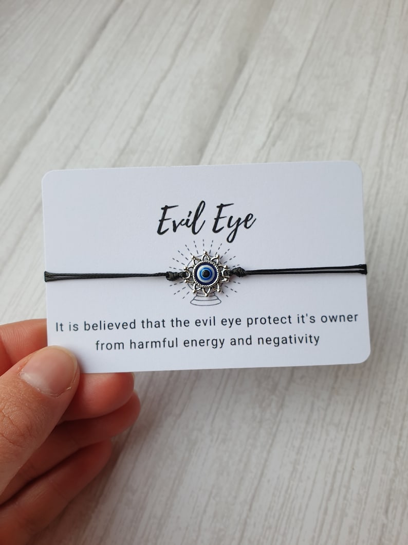 May include: A black string bracelet with a silver charm featuring a blue evil eye. The card reads "Evil Eye" and "It is believed that the evil eye protect it's owner from harmful energy and negativity."