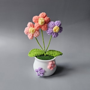 May include: A small crocheted flower arrangement in a white pot. The flowers are pink, purple, and yellow. The pot has a green base and is decorated with two small crocheted flowers.