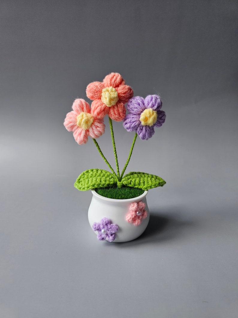 May include: A small crocheted flower arrangement in a white pot. The flowers are pink, purple, and yellow. The pot has a green base and is decorated with two small crocheted flowers.