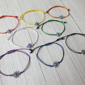 May include: Eight colorful string bracelets with a silver evil eye charm. The bracelets are red, yellow, orange, green, white, blue, purple, and black.