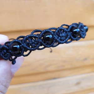 May include: Black macrame bracelet with five glossy, round black beads. The bracelet has an intricate, open weave design. The bracelet is a dark, neutral colour, suitable for everyday wear. The bracelet's length is approximately 18 cm.