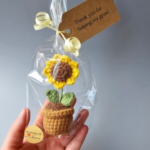 May include: A small crocheted sunflower plant in a brown pot with green leaves. The plant is packaged in a clear plastic bag with a brown gift tag that says "Thank you for helping me grow!" and a small brown sticker that says "handmade with love".