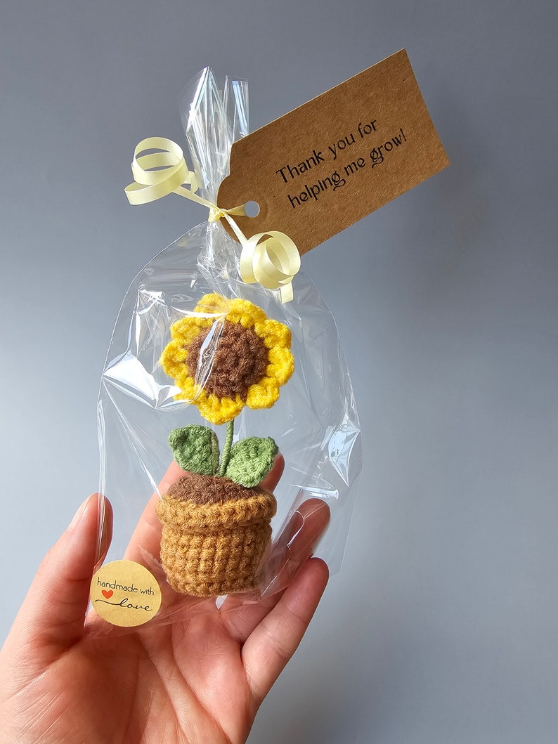 May include: A small crocheted sunflower plant in a brown pot with green leaves. The plant is packaged in a clear plastic bag with a brown gift tag that says "Thank you for helping me grow!" and a small brown sticker that says "handmade with love".