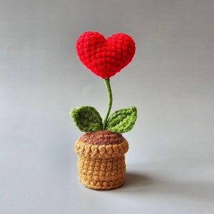 May include: A crocheted heart-shaped flower in a brown pot with green leaves. The flower is red and the pot is brown.